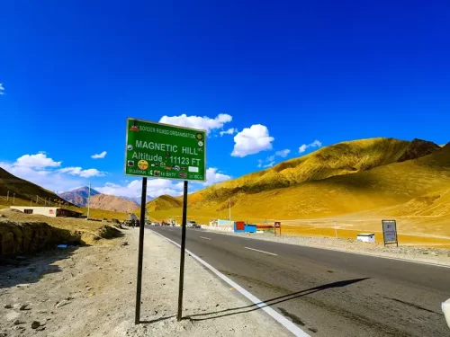 Magnetic Hill sign at Ladakh BRO during clear skies, featuring altitude 11312 ft board yellow mountains highway, perfect adventure Ladakh tour package. 