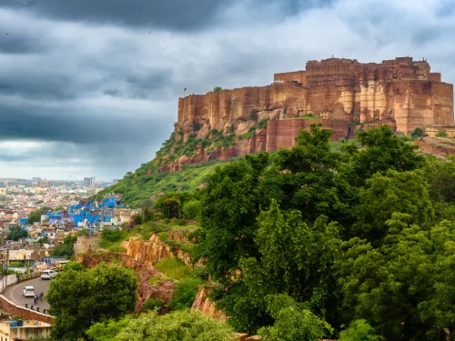Mehrangarh Fort dramatic vista during cloudy day, featuring imposing pink ramparts atop rocky hill overlooking Jodhpur blue cityscape, perfect cultural heritage Rajasthan tour packages.