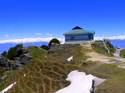 Mahasu Peak in Kufri featuring a small hilltop building with green roof, winding mountain road, grassy slopes with patches of snow, and panoramic snow-capped Himalayan ranges under a clear blue sky.