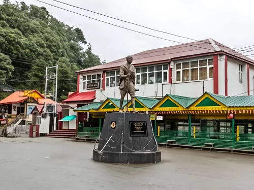 Mall Road in Dalhousie featuring a central statue of Mahatma Gandhi on a pedestal, surrounded by colorful shopfronts and colonial-style buildings under an overcast sky.