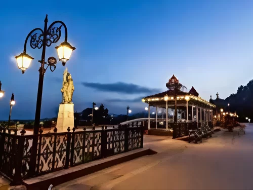 Indira Gandhi statue at Mall Road Shimla during twilight, featuring lit bandstand, vintage lamps, railing, hills, perfect evening stroll, Himachal Pradesh tour packages.