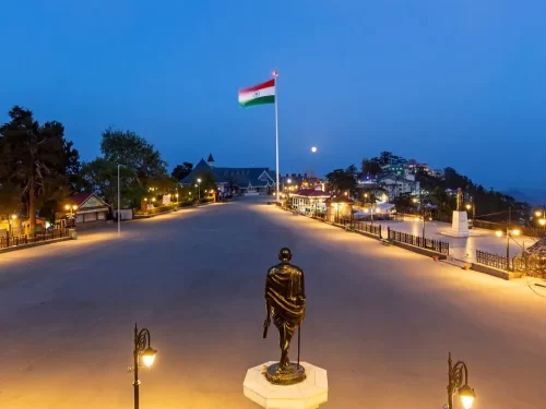 Gandhi Statue at Mall Road Shimla during twilight, featuring Indian flag, colonial buildings, lamps, hills, perfect evening stroll, Himachal Pradesh tour packages.