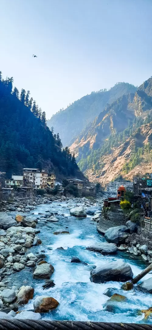 Himachal Pradesh Tour Package featuring scenic river flowing through rocky valley with mountain village and pine-covered hills in the backdrop.