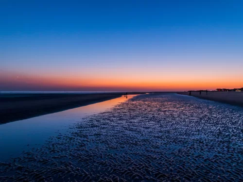 Mandvi Beach sunset at Kutch Gujarat during twilight, featuring distant silhouettes, blue-pink skies, water reflections on white sands and Arabian Sea waves, perfect romantic experience with Gujarat tour packages.