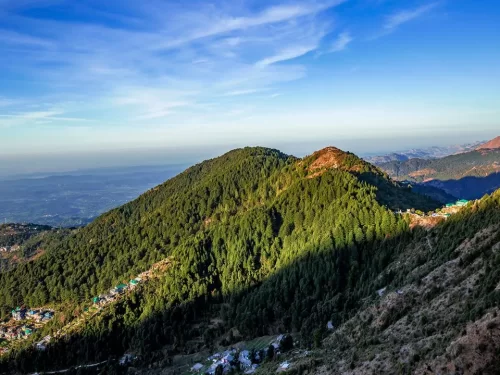 McLeod Ganj Dharamshala Himachal Pradesh during golden hour sunset lighting partly cloudy skies, featuring panoramic pine forested hills scattered buildings distant valley mountain ranges backdrop, perfect cultural experience Himachal tour package.