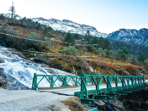 Green bridge over Khanda waterfall at Katao Lachung Sikkim amid prayer flags and snow peaks, perfect adventure North Sikkim tour package.