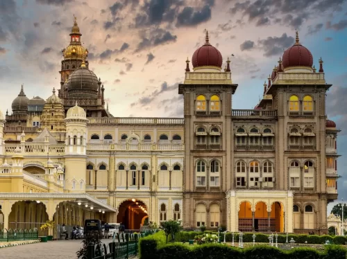 Mysore Palace illuminated at sunset Mysuru Karnataka, featuring golden domes red towers arched facade gardens clouds, perfect royal romantic Karnataka tour package.