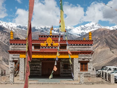 Nako Monastery Spiti Valley during clear blue skies, featuring red yellow building prayer flags snow-capped mountains vehicles courtyard, perfect spiritual experience Himachal Pradesh tour package.