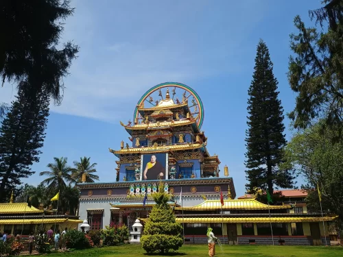 Golden Temple at Namdroling Monastery Bylakuppe during sunny day, featuring multi-tiered roofs, Dalai Lama portrait, prayer wheel, trees, perfect cultural experience Karnataka tour packages.