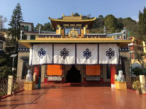 Nechung Monastery in Dharamshala featuring a traditional Tibetan-style temple with golden roof details, colorful facade, lion statues at the entrance, and hillside buildings in the background.
