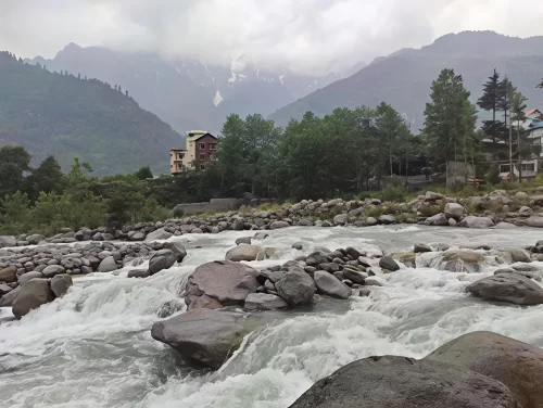 Rushing river at Nehru Kund Manali during misty weather, featuring rocky banks pine forests and snow peaks, perfect nature experience Himachal Pradesh tour package.