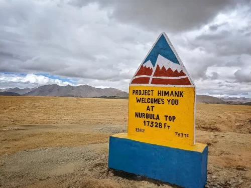 Nurbula Top (Nurbula Pass) Ladakh signboard Project Himank welcomes 17328 ft Nubra 1753 ft hazy mountains barren plateau gravel road, perfect high-altitude adventure Ladakh tour package.