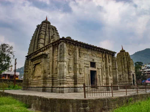 Historic Panchavaktra Temple featuring intricate stone carvings, tall shikhara towers with red flags, and ancient architectural detailing set against a cloudy sky and surrounding hills.