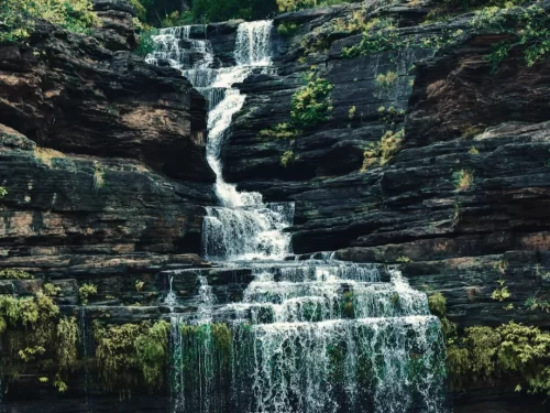 Pandav Falls in Madhya Pradesh, a multi-tiered waterfall cascading over dark layered rock formations surrounded by lush greenery, a scenic natural attraction featured in Madhya Pradesh tour packages.