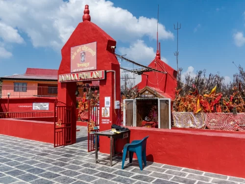 Pohlani Mata Temple in Dalhousie featuring bright red temple structures, entrance gate with “Jai Maa Pohlani” signage, prayer flags, tridents, and shrine area under a blue sky.