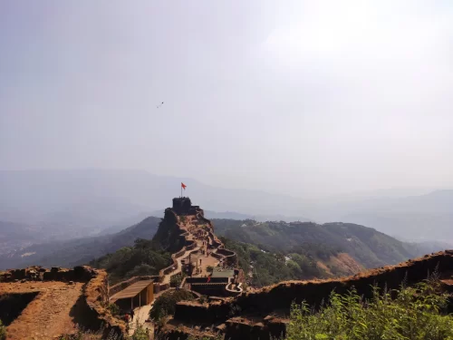 Pratapgad Fort Mahabaleshwar elevated trails, featuring winding ramparts red saffron flag atop peak misty valleys green hills hazy skies, perfect Maharashtra tour packages.