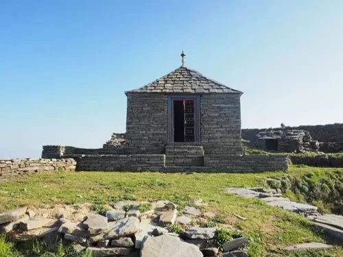 Stone structure of Raghupur Fort in Jibhi featuring a small historic watchtower-style building with slate roof, surrounded by grassy hilltop and ancient fort ruins under a clear blue sky.
