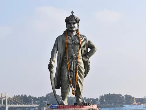 Raja Bhoj statue at Bhopal Upper Lake during clear daylight, featuring sword garlands river bridge backdrop, perfect cultural Madhya Pradesh tour package.