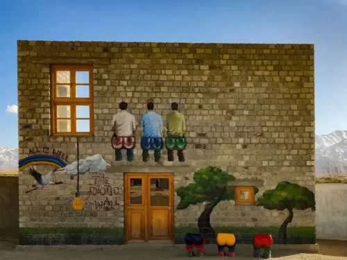 Rancho School mural at Ladakh during clear skies, featuring 3 Idiots boys rainbow trees door mountains backdrop, perfect adventure Ladakh tour package.