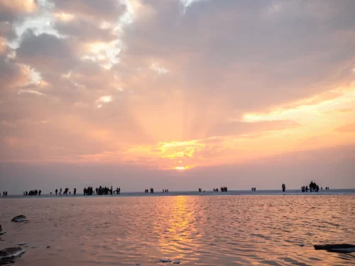 Rann of Kutch sunset during golden hour, featuring silhouetted tourist crowds, radiant sun rays, pink-orange skies and vast white salt desert, perfect adventure experience with Gujarat tour packages.