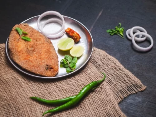 Rava Fried Fish is a crispy semolina-coated fish fillet, shallow fried to golden perfection and served with onion rings, lime wedges, and chutney for a flavorful coastal delight.
