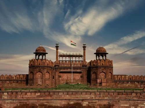 Red Fort Lahori Gate at Delhi on partly cloudy day, featuring Indian flag and red sandstone towers, perfect heritage Delhi tour package.