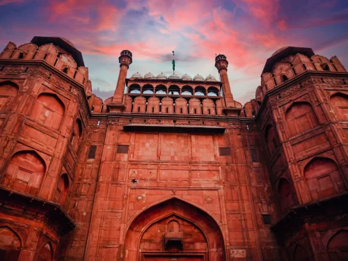 Red Fort Lahori Gate at Delhi during sunset, featuring Indian flag and red sandstone towers, perfect heritage Delhi tour package.