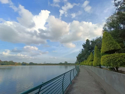 Sankey Tank Bangalore lakeside walkway during sunny day with clouds, featuring green hedges, trees and calm water, perfect Karnataka tour package.