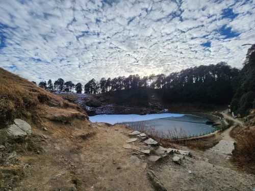 Icy Serolsar Lake Jibhi during winter cloudy skies, featuring pine forests, trek path, rocky shores, perfect serene Himachal Pradesh tour package.