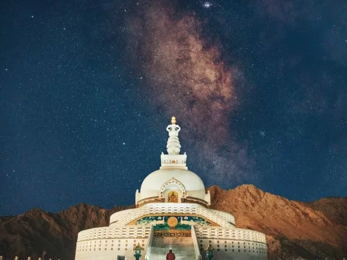 Shanti Stupa Leh Ladakh beneath starry Milky Way night sky, featuring white stupa golden spire dome, colorful base stairs, silhouetted mountain backdrop, visitors, perfect astrophotography spiritual stargazing experience with Leh Ladakh night sightseeing 