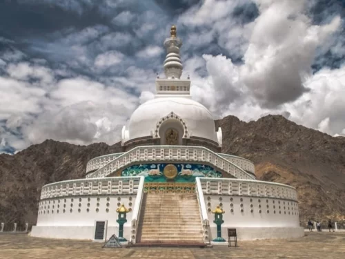 Shanti Stupa Leh Ladakh under partly cloudy sky, featuring white-domed Buddhist chorten with golden spire, colorful base murals, steep stairs, rugged mountains backdrop, perfect peace panoramic experience with Leh city sightseeing tour package. 