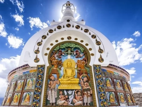 Close-up of Shanti Stupa Leh Ladakh entrance under partly cloudy sky, featuring golden Buddha statue, vibrant murals of deities, white stupa dome with spire, railings, perfect Buddhist heritage experience with Leh Ladakh sightseeing tour package. 