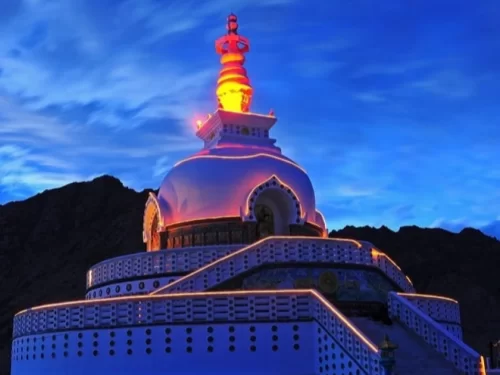 Illuminated Shanti Stupa Leh Ladakh at night with pink blue twilight sky, featuring glowing white dome stupa golden spire, colorful railings, rugged mountain backdrop, perfect spiritual night view experience with Leh Ladakh sightseeing tour package.