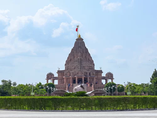 Shree Khodaldham Temple at Kagvad Rajkot during sunny partly cloudy skies, featuring towering shikhara, saffron flag, intricate pink stone carvings and lush gardens, perfect spiritual experience with Gujarat tour packages.