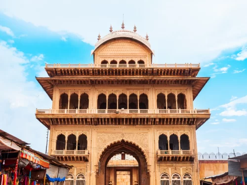 Ornate haveli-style gate of Sri Rangji Mandir during partly cloudy daytime, featuring multi-tiered sandstone architecture, bustling market surroundings, perfect spiritual heritage experience with Vrindavan tour package