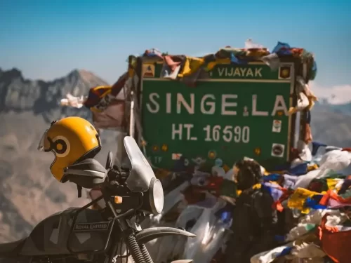 Royal Enfield biker yellow helmet at Singe La Pass HT 16590 ft Lahaul Spiti sunny day, prayer flags snow peaks; perfect Ladakh Manali tour package.