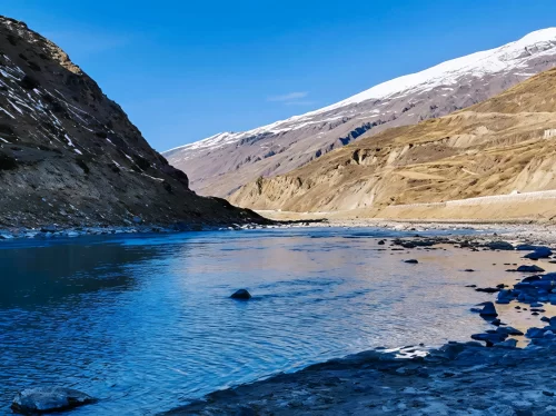 Sissu Valley serene turquoise river meandering through barren brown canyon walls with light snow patches on distant peaks under clear blue skies, rocky shores, perfect Himachal Pradesh adventure tour package.