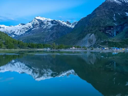 Sissu Lake mirror reflection of snow-capped peaks, cascading waterfall, colorful village huts with green trees along deep blue waters under twilight blue sky, perfect Himachal Pradesh tour package.
