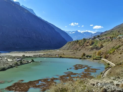 Sissu Lake vibrant turquoise pond amid barren rocky valley with terraced autumn foliage villages, towering gray peaks under blue sky with clouds, path encircling algae patches, perfect Himachal Pradesh tour package.