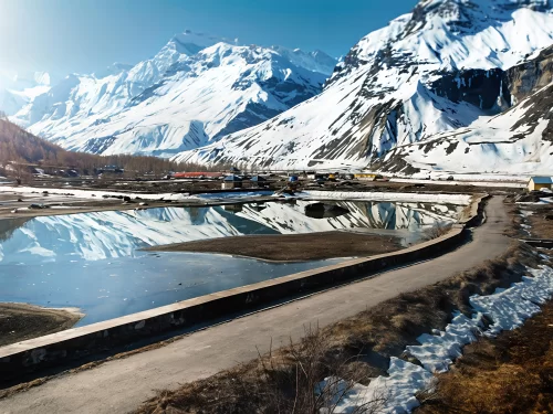 Sissu Lake in Lahaul Valley during winter daylight, featuring icy blue waters with perfect reflections of snow-capped Himalayan peaks, scattered village huts and concrete path edged by snow, perfect Himachal Pradesh tour package.