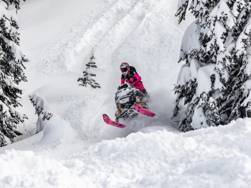 Snow scooter ride through deep powder snow, featuring a rider in bright pink winter gear navigating between snow-covered trees in a mountainous landscape.