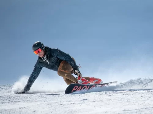 Snowboarding activity with a rider carving through fresh snow on a mountain slope, wearing a helmet and goggles while kicking up powder against a clear blue sky.