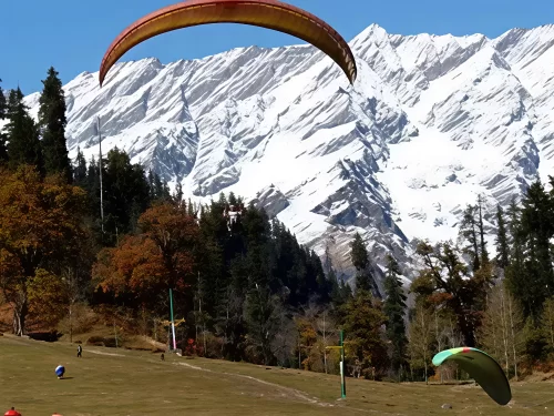 Paragliding adventure at Solang Valley Manali during sunny skies, featuring orange green gliders over snowy peaks pine forests and meadows, perfect adventure experience Himachal Pradesh tour package.