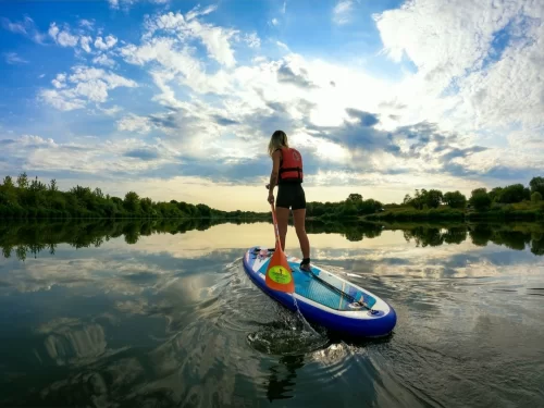 Stand-up paddleboarding activity with a person wearing a life jacket paddling on a calm river, surrounded by lush greenery and dramatic clouds reflecting on the water during sunset.