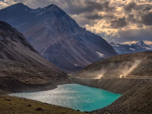 Dramatic Suraj Tal high-altitude lake below Baralacha La Pass on Manali Leh Highway Himachal Pradesh during golden hour sunset, featuring turquoise waters rugged mountains dust trail truck dramatic clouds snowy peaks, perfect road trip adventure experienc