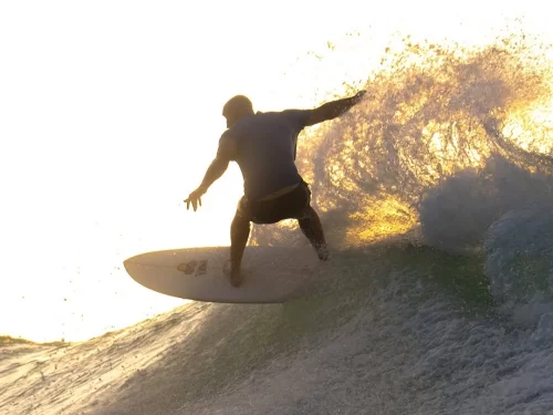 Surfing activity with a surfer riding a powerful ocean wave at sunset, silhouetted against golden light with water splashing dramatically around the surfboard.