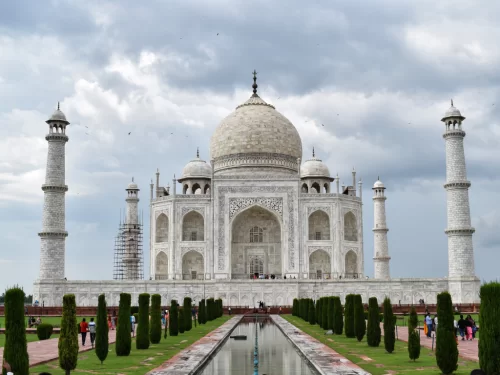 Classic Taj Mahal front facade Agra Uttar Pradesh under cloudy skies with tourists, reflecting pool, cypress trees, white marble mausoleum, four minarets, perfect UNESCO heritage India tour package. 