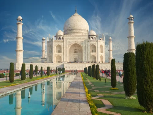 Taj Mahal Agra Uttar Pradesh with perfect reflection in reflecting pool, tourists along cypress avenue, white marble mausoleum, four minarets, partly cloudy blue skies, perfect UNESCO heritage India tour package.