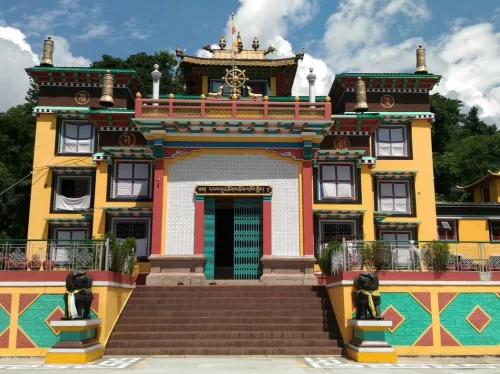 Front view of Tashi Thongyang Monastery featuring a vibrant yellow and red Tibetan-style facade, central entrance with decorative doorway, prayer symbols, rooftop ornaments, and lion statues at the staircase under a blue sky.