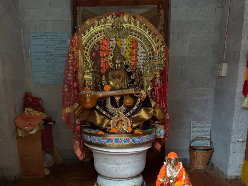 Teetwal Village Sharda Devi idol shrine during indoor lighting Kashmir, featuring golden veena garlands red drapes pedestal small Hanuman statue stone walls, perfect Kashmir tour packages.